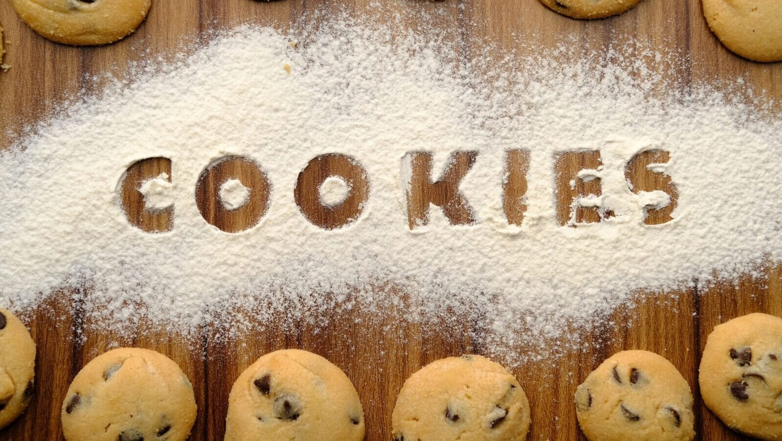 Freshly baked chocolate chip cookies arranged on flour-dusted wooden table.